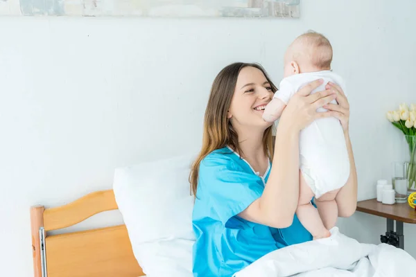 Sonriente y atractiva madre sosteniendo a su hijo en el hospital - foto de stock