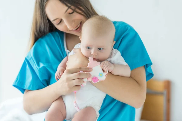 Attraktive und lächelnde Mutter hält ihr Kind mit Spielzeug im Krankenhaus — Stockfoto