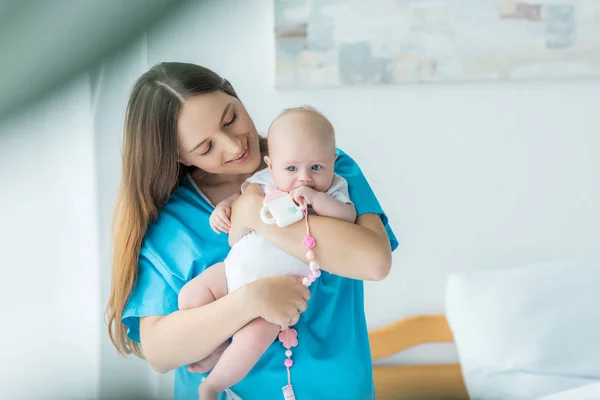 Привлекательная и улыбающаяся мать держит своего ребенка с игрушкой в больнице — стоковое фото