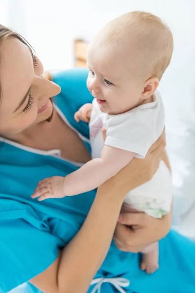 Atractiva y sonriente madre sosteniendo a su hijo en el hospital - foto de stock