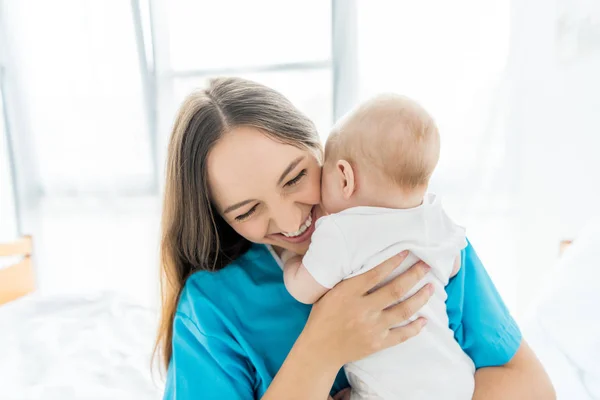 Atractiva y sonriente madre sosteniendo a su hijo en el hospital - foto de stock