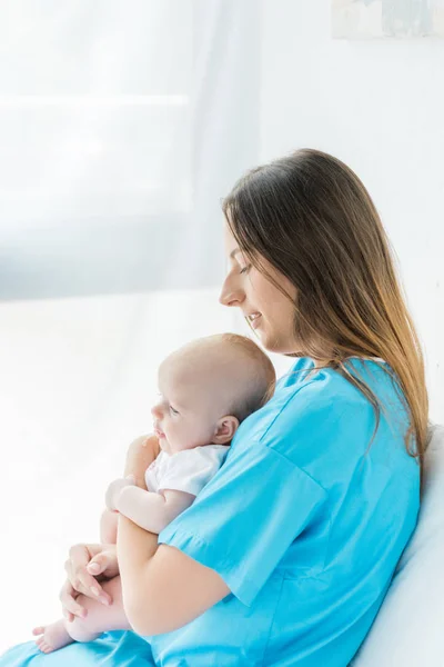 Vue latérale de belle et jeune mère tenant son enfant à l'hôpital — Photo de stock