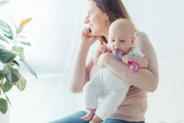 Attractive and smiling mother holding her child and talking on smartphone in apartment — Stock Photo