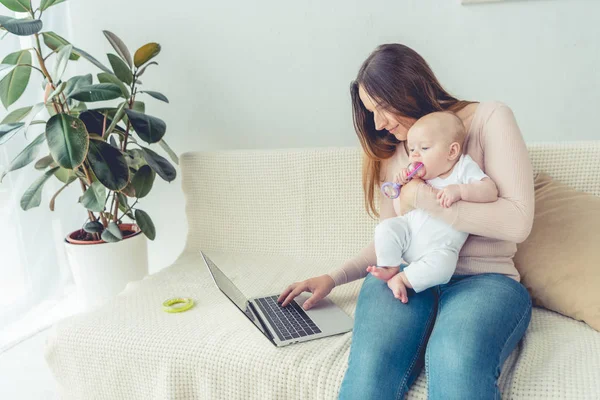 Atractiva madre sosteniendo a su hijo y utilizando el ordenador portátil en el apartamento - foto de stock