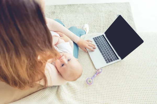 Cropped view of mother holding her child and using laptop in apartment — Stock Photo