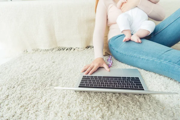Обрезанный вид матери, держащей ребенка и с помощью ноутбука в квартире — стоковое фото