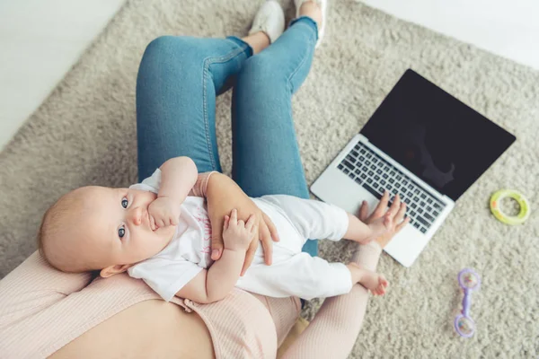 Vista ritagliata della madre che tiene in braccio il suo bambino e utilizza il computer portatile in appartamento — Foto stock