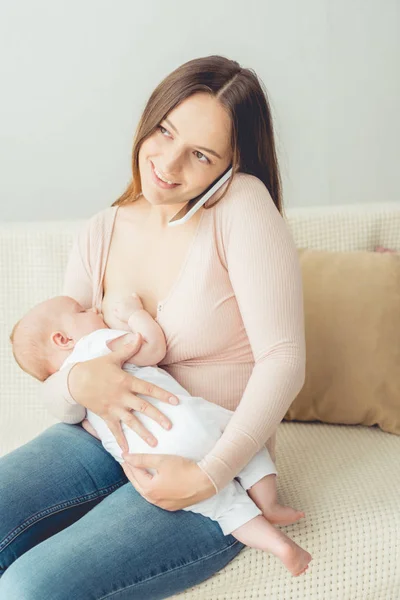 Mãe atraente amamentando seu filho e falando no smartphone no apartamento — Fotografia de Stock