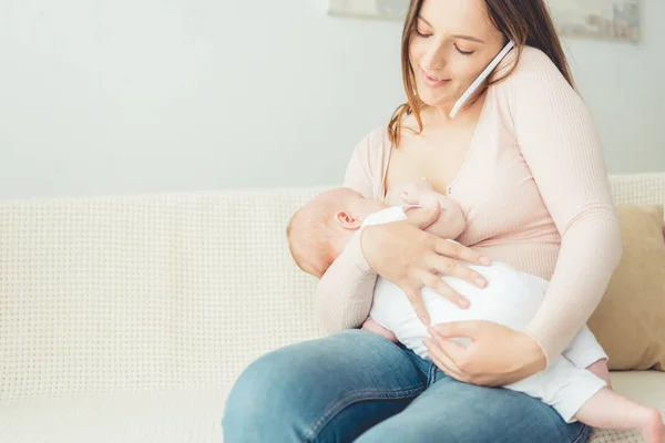 Attractive mother breastfeeding her child and talking on smartphone in apartment — Stock Photo