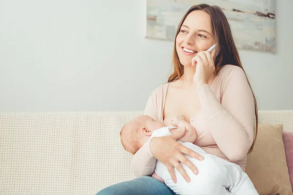 Attractive mother breastfeeding her child and talking on smartphone in apartment — Stock Photo