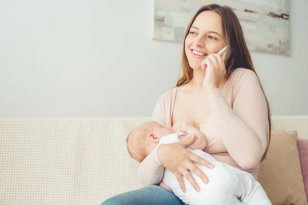 Mãe atraente amamentando seu filho e falando no smartphone no apartamento — Fotografia de Stock