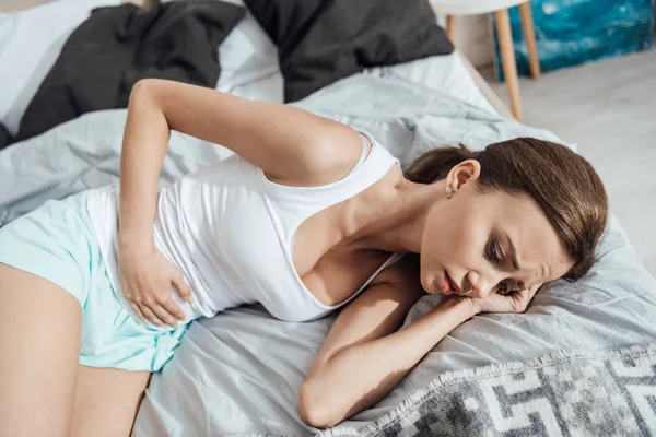 Aufgebrachte junge Frau liegt auf Bett und berührt Bauch — Stockfoto