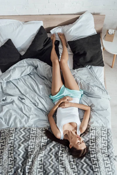 Vista superior de la mujer joven molesta acostada en la cama y tocando el vientre - foto de stock