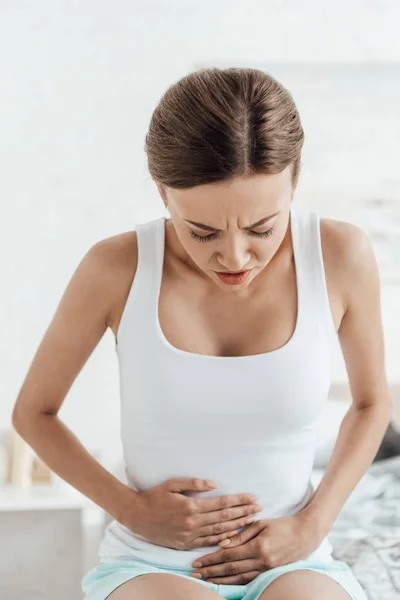 Aufgebrachte junge Frau sitzt auf Bett und berührt Bauch — Stockfoto