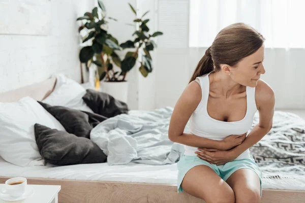 Aufgebrachte junge Frau sitzt auf Bett und berührt Bauch — Stockfoto