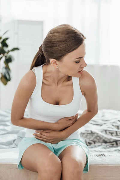 Aufgebrachte junge Frau sitzt auf Bett und berührt Bauch — Stockfoto