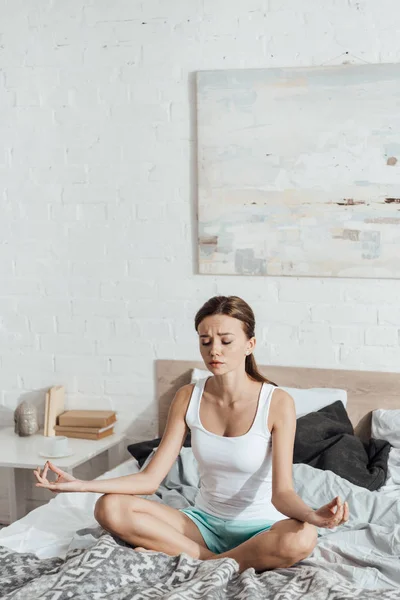 Aufgebrachte junge Frau sitzt mit geschlossenen Augen in Lotus-Pose auf Bett — Stockfoto
