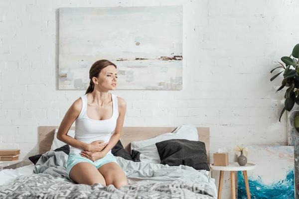 Напряженная молодая женщина сидит на кровати и трогает живот — стоковое фото