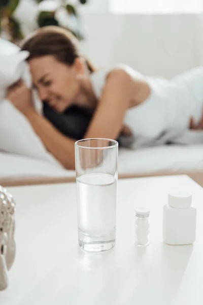 Selective focus of suffering woman in bed and pills with glass of water on foreground — Stock Photo