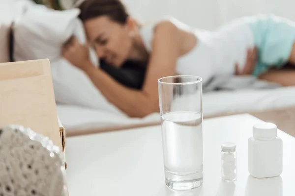 Selektiver Fokus der leidenden Frau im Bett und Tabletten mit Glas Wasser im Vordergrund — Stockfoto