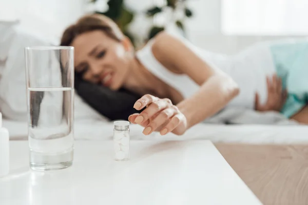 Selektiver Fokus der leidenden Frau im Bett und Tabletten mit Glas Wasser im Vordergrund — Stockfoto