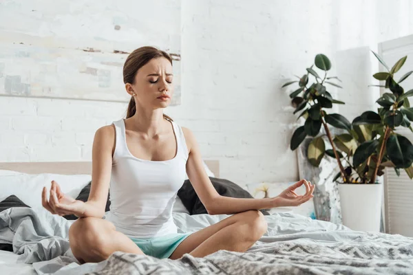 Upset young woman sitting on bed in lotus pose with closed eyes — Stock Photo