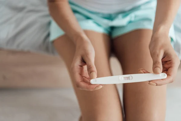 Partial view of young woman sitting on bed and holding pregnancy test — Stock Photo