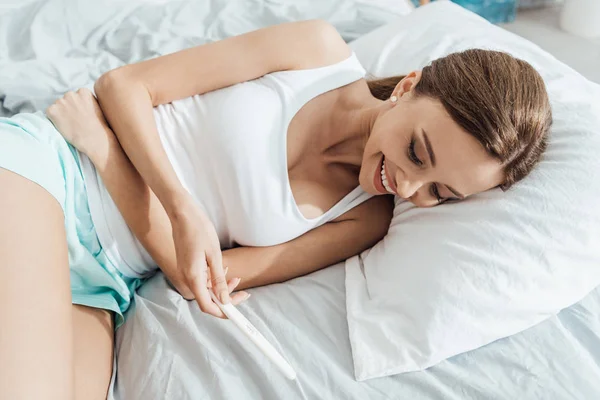 Happy young woman lying in bed and holding pregnancy test — Stock Photo