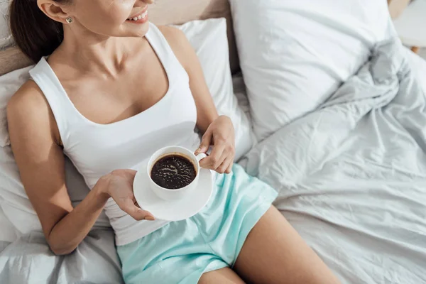 Vista parziale di giovane donna sdraiata a letto e con in mano una tazza di caffè — Foto stock