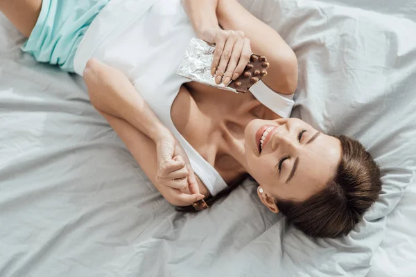 Вид зверху усміхненої молодої жінки, що їсть шоколад з горіхами в ліжку — стокове фото