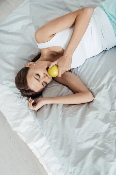 Над головой вид грустной молодой женщины, которая ест зеленое яблоко в постели — стоковое фото