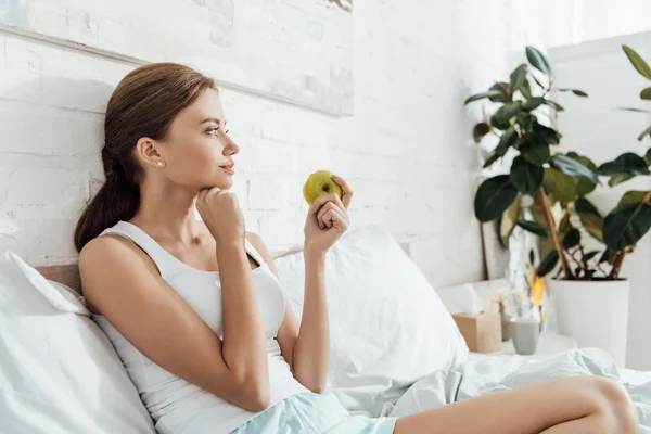 Attractive young woman lying in bed and holding green apple — Stock Photo