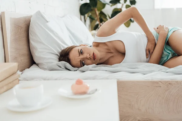 Leidende Frau liegt im Bett, berührt Bauch und schaut auf Cupcake — Stockfoto