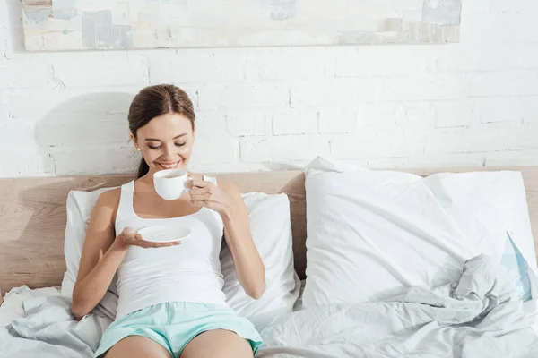 Улыбающаяся молодая женщина лежит в постели и пьет кофе — стоковое фото