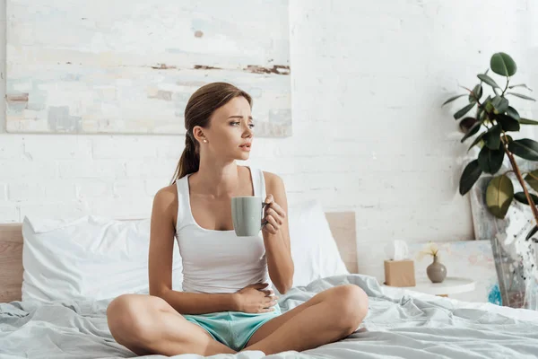 Stressé jeune femme assise sur le lit, toucher le ventre et tenant une tasse de thé — Photo de stock