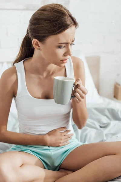Напряженная молодая женщина сидит на кровати, касаясь живота и держа чашку чая — стоковое фото