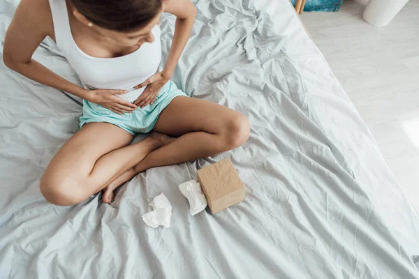 Overhead view of stressed young woman sitting on bed and touching belly — Stock Photo