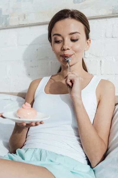 Весела молода жінка лежить в ліжку і їсть кекс — стокове фото