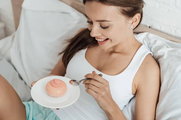 Alegre joven mujer acostada en la cama y comiendo cupcake - foto de stock