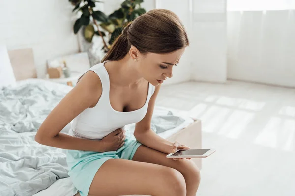 Triste joven mujer tocando el vientre y el uso de teléfono inteligente en el dormitorio - foto de stock
