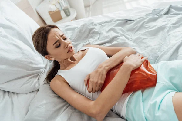 Suffering young woman lying in bed and holding warmer — Stock Photo