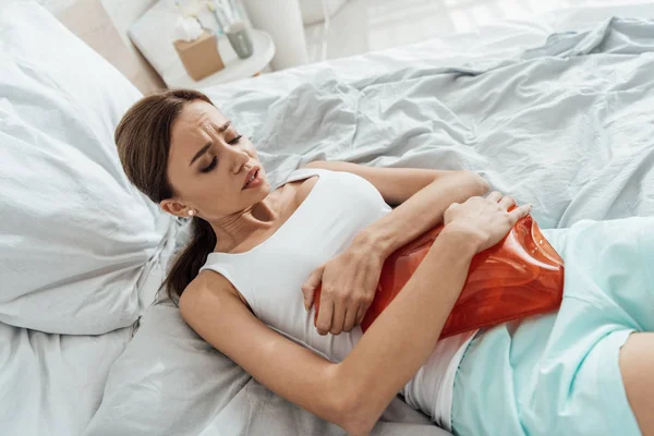 Souffrance jeune femme couché dans le lit et tenant plus chaud — Photo de stock