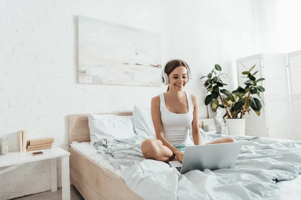 Smiling young woman listening music in headphones and using laptop on bed — Stock Photo