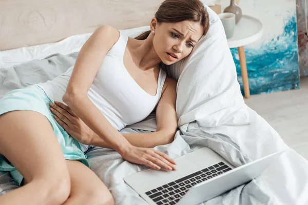 Mujer joven molesta acostada en la cama con el ordenador portátil y tocando el vientre - foto de stock