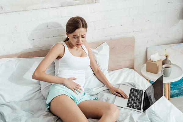 Upset young woman lying on bed with laptop and touching belly — Stock Photo