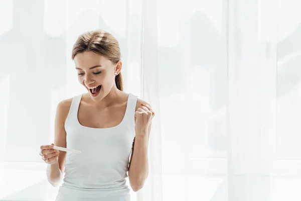 Sonriente joven mujer sosteniendo prueba de embarazo y mostrando sí gesto en casa - foto de stock