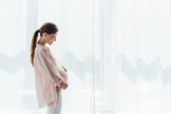 Seitenansicht einer glücklichen Schwangeren, die lächelt und ihren Bauch berührt — Stockfoto