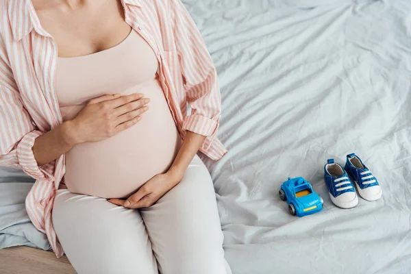 Vista parziale della donna incinta con auto giocattolo e scarpe per bambini seduta sul letto e pancia toccante — Foto stock