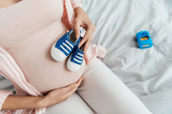 Teilansicht einer schwangeren Frau mit Spielzeugauto und Babyschuhen, die auf dem Bett sitzt und Bauch berührt — Stockfoto