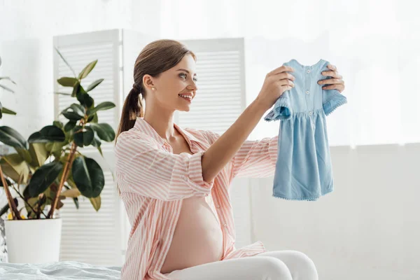 Happy smiling pregnant woman sitting on bed and holding baby clothes — Stock Photo